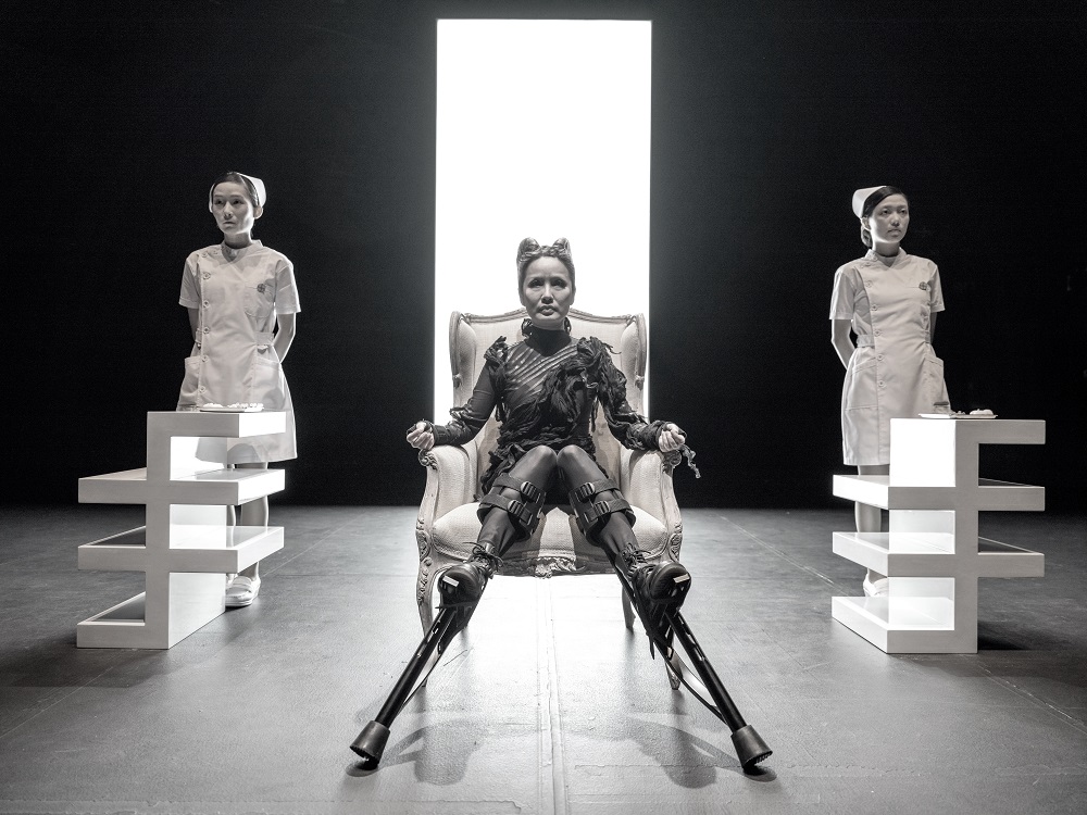 Black and white photo of a woman sits in a chair wearing black ruffled material, futuristically, fashionably cut. She wears stilts on her feet. The chair is ornate, in a palatial style, Edwardian possibly. Two nurses stand either side of the seated woman, stood behind two squared of dollar signs in white, which come to the tops of their thighs. Directly behind the woman is a bright white open doorway almost ceiling height.