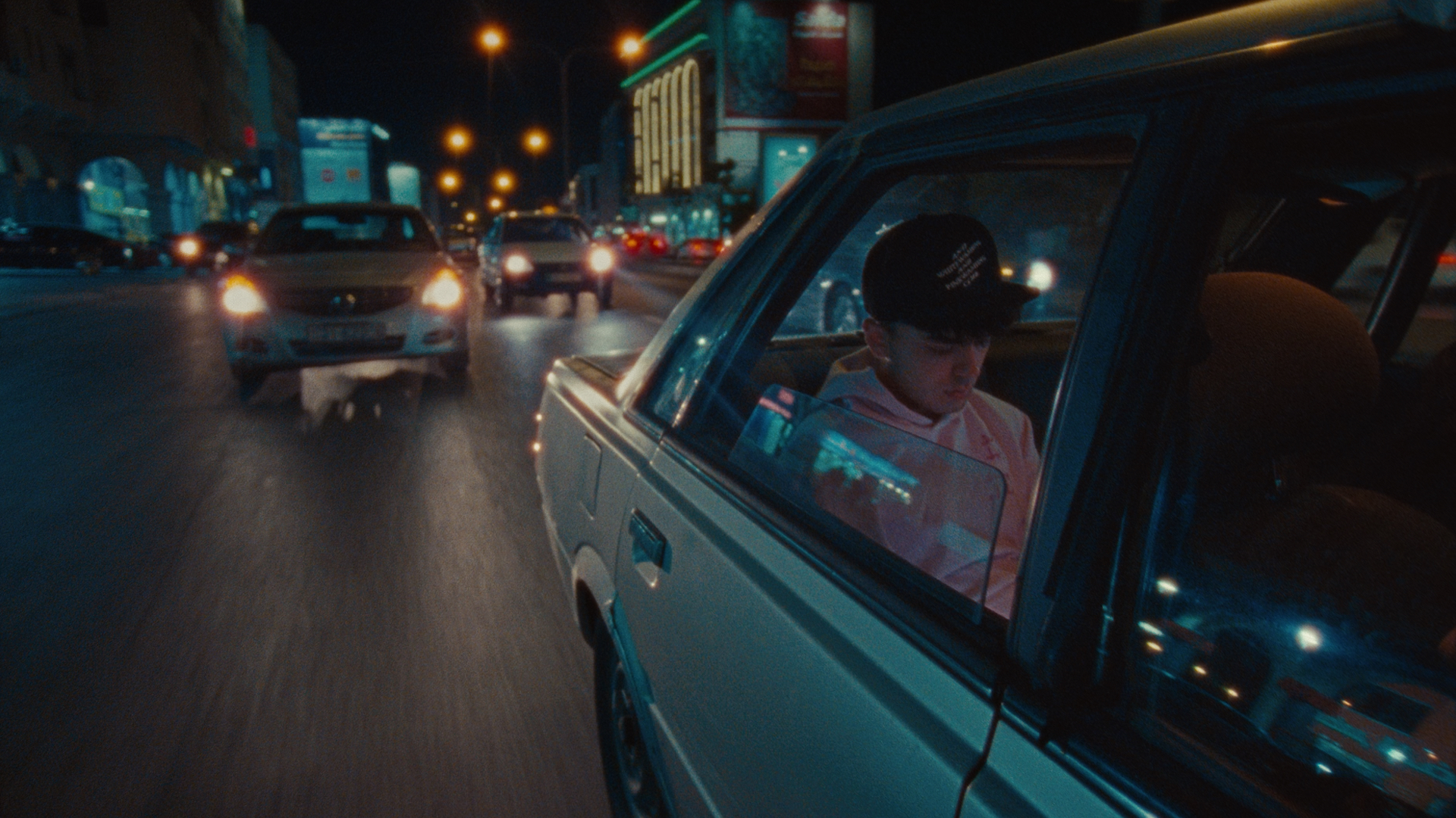 A young man sits in the back seat of a car with the window open, he wears a black cap and pink hooded sweatshirt. The car is driving down a busy street at night. Other cars follow behind with their lights on. Lit up buildings and streetlights overhead can be seen behind the car.