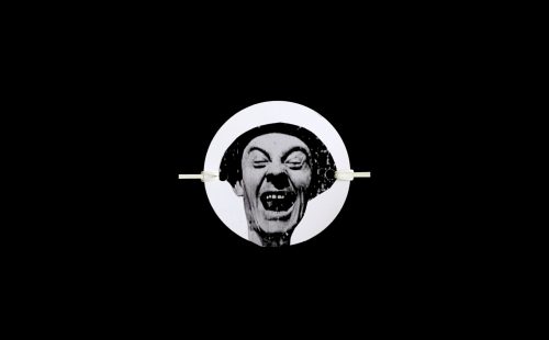 Black and white picture of a man in a white circle laughing wildly waering a black flat cap.