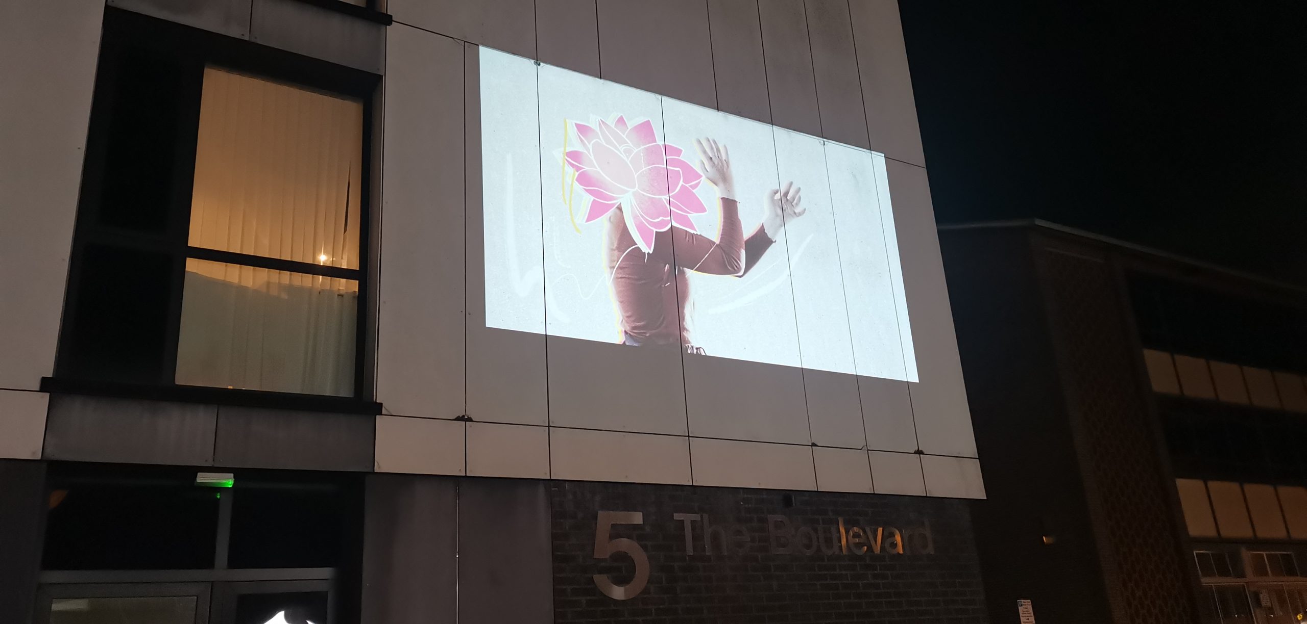 Projection onto a white residential tower block of a woman holding her hands in front of her with an illustration of a pink carnation covering her face and head over a white background.