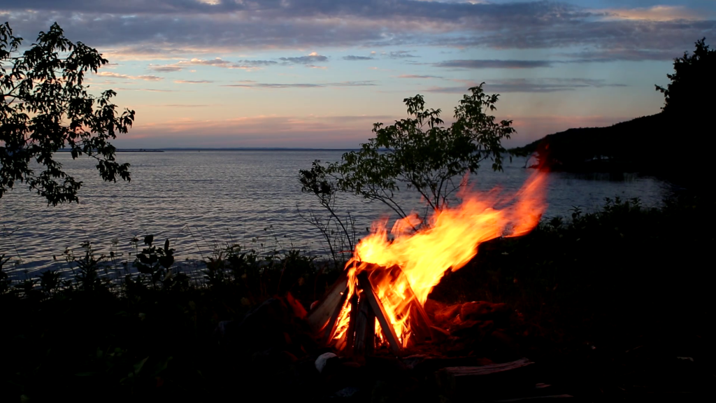 A bonfire on the beach of a large lake at sunrise, blue skies with an orange morning glow in the distance.