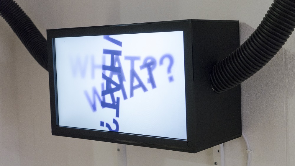 Image of a black rectangular lightbox with a white screen has the word what with a question mark written 3 times on it, black pipes leave the box on the left and right sides.