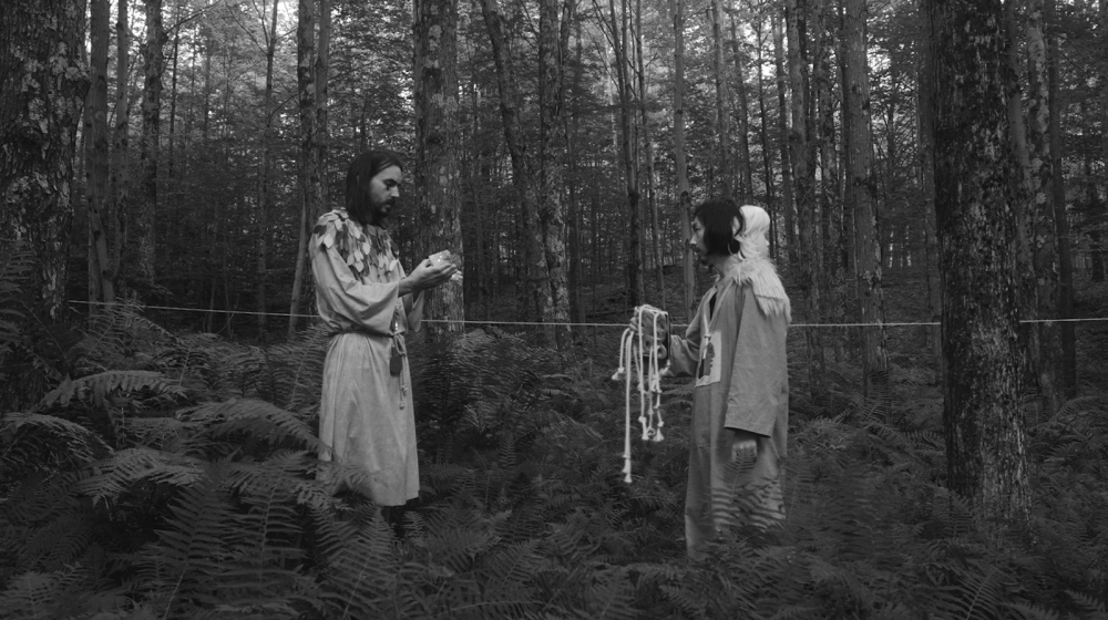 You are looking at a black-and-white image. The scene is in a wood, you will see trees at the back and tall grass on the ground that reaches the waist of an adult. Two men standing in the center of the image facing each other. They both wearing the ancient look with robes. The man on the left is taller with four eyes, holding a crystal cube. The man on the right has another head attached to his head with white hair. He is holding a book with a rope on a book cover. It looks like the man on the right is talking to the man on the left.
