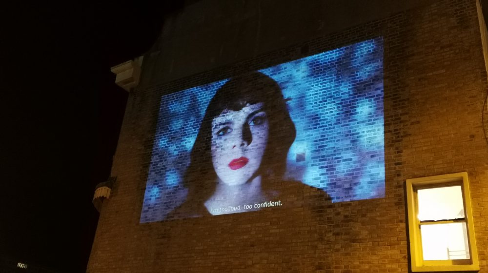 Large projection onto the brick wall of a regency townhouse of a woman with black hait and bright red lipstick and a shimmering blue background.