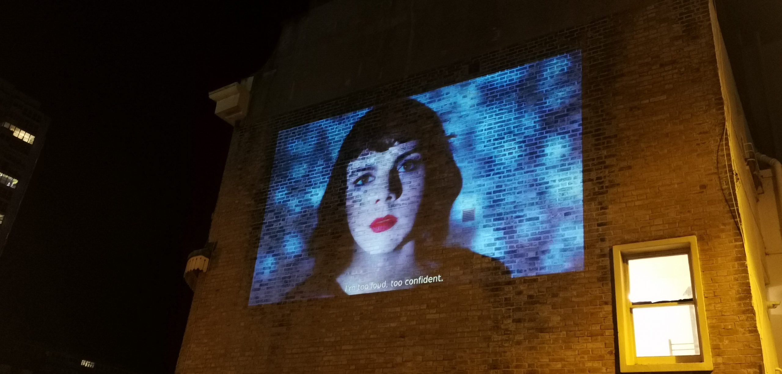 Large projection onto the brick wall of a regency townhouse of a woman with black hait and bright red lipstick and a shimmering blue background.