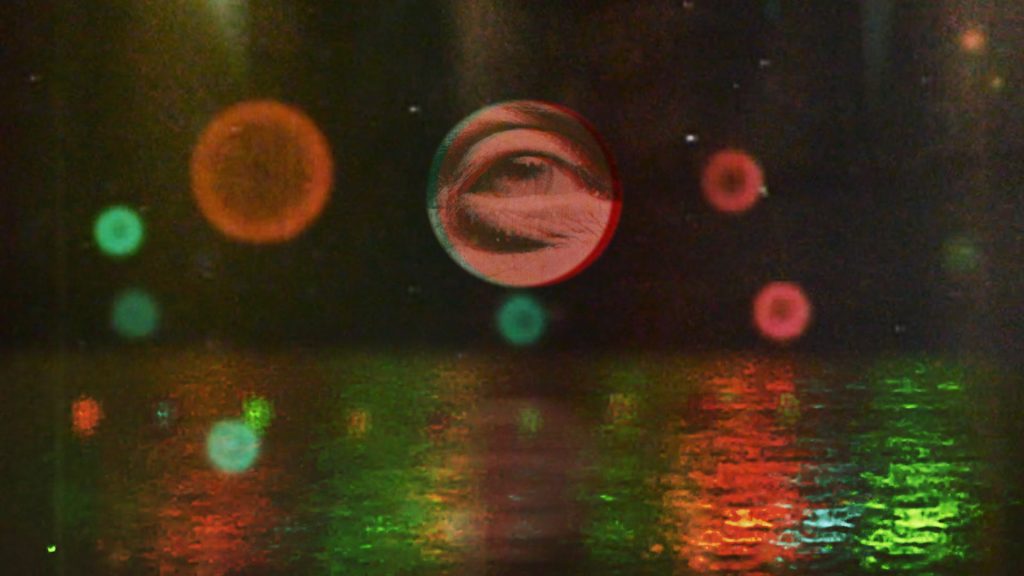 An eye peers out from the centre of a circle on a black background with red and green circles floating over shimmering water lit in red and green.
