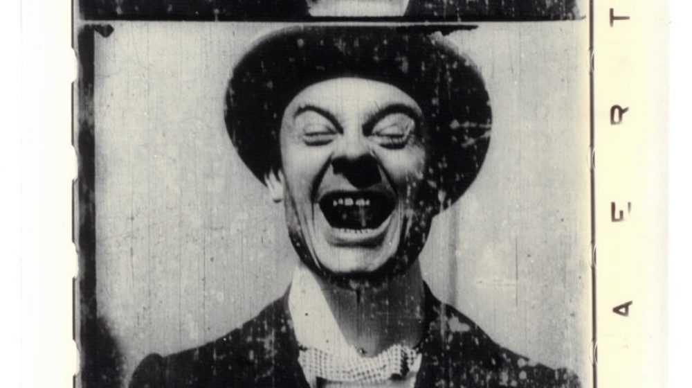 A black and white still image from a film shows a man wearing a round brimmed hat, his mouth is opening, teeth showing, his eyes are closed and eyebrows are arched highly.
