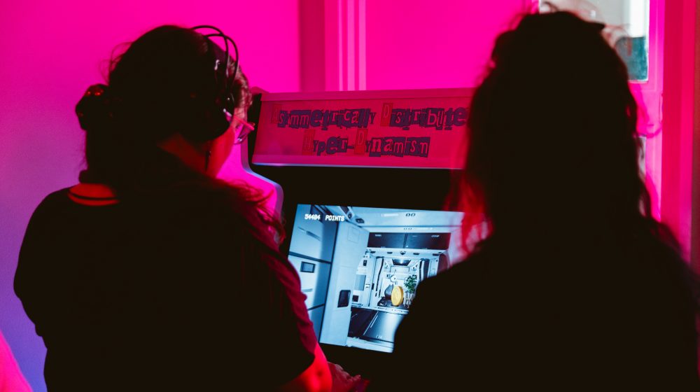 2 people stand in front of a pink neon lit videogame arcade. One person is playing the game which is titled on top Asymmetrically Desirable Hyper Dynamism.