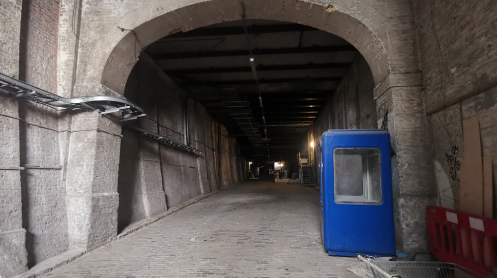 An image of a deep tunnel madeof brick with an archway at the beginning. The floor is made of grey cobbles.