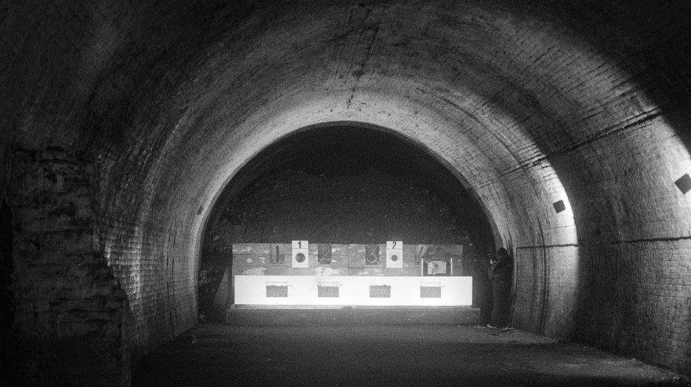 A dimly lit underground tunnel with a curved brick ceiling. At the far end, illuminated targets are arranged on a stand, numbered '1' and '2.' A person is standing to the right of the targets, partially in shadow. The scene has a stark, atmospheric quality with contrasting light and dark areas, emphasising the tunnel's depth and texture.