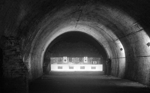 A dimly lit underground tunnel with a curved brick ceiling. At the far end, illuminated targets are arranged on a stand, numbered '1' and '2.' A person is standing to the right of the targets, partially in shadow. The scene has a stark, atmospheric quality with contrasting light and dark areas, emphasising the tunnel's depth and texture.