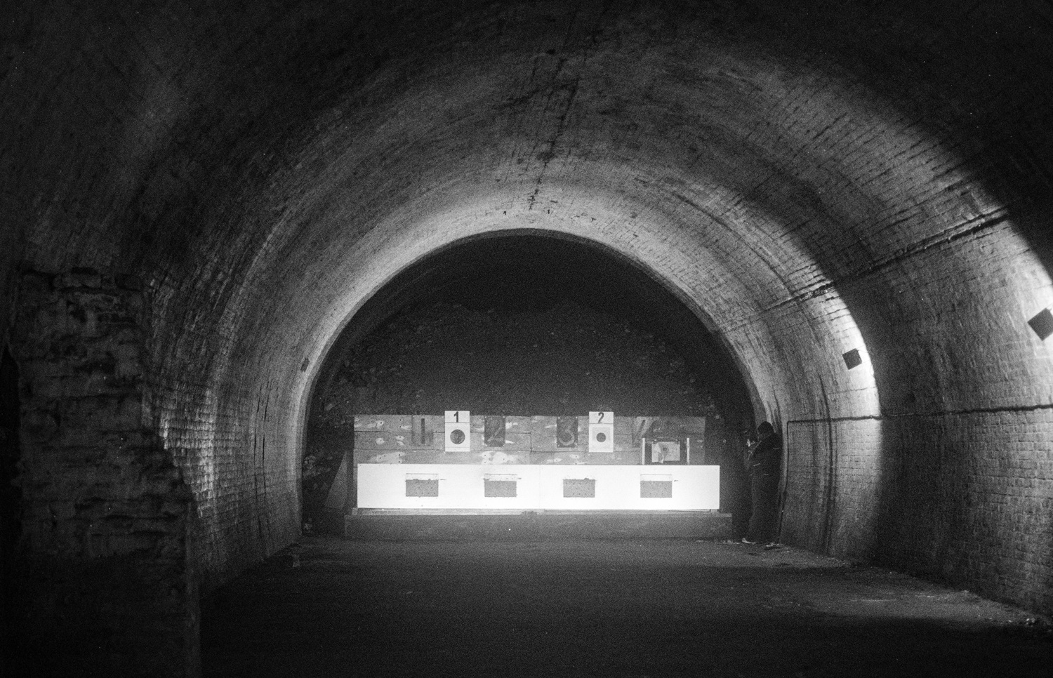 A dimly lit underground tunnel with a curved brick ceiling. At the far end, illuminated targets are arranged on a stand, numbered '1' and '2.' A person is standing to the right of the targets, partially in shadow. The scene has a stark, atmospheric quality with contrasting light and dark areas, emphasising the tunnel's depth and texture.