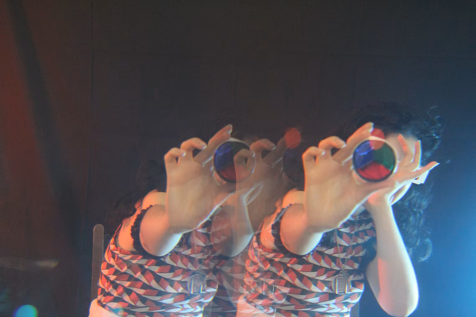 A person with curly dark hair holds a circular object with red, green, and blue segments in front of their face, creating a colourful lens effect. They are wearing a patterned red, black, and white top and are set against a dark background with a bright light source to the right. The image has a double exposure or motion blur effect, making it appear as though the person is duplicated.