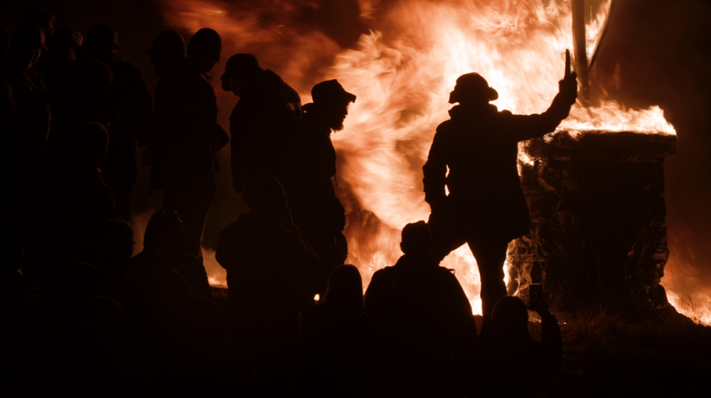 A silhouetted crowd of people gathered around a large bonfire at night, with flames dramatically illuminating the scene. One person in the foreground holds up a torch or object beside a burning stone structure, creating a powerful and intense visual contrast between shadow and fire.