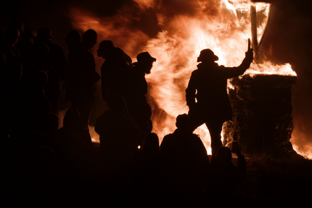 A silhouetted crowd of people gathered around a large bonfire at night, with flames dramatically illuminating the scene. One person in the foreground holds up a torch or object beside a burning stone structure, creating a powerful and intense visual contrast between shadow and fire.