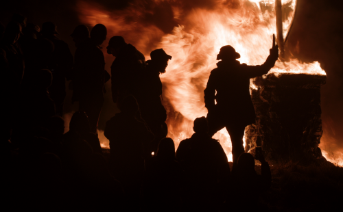 A silhouetted crowd of people gathered around a large bonfire at night, with flames dramatically illuminating the scene. One person in the foreground holds up a torch or object beside a burning stone structure, creating a powerful and intense visual contrast between shadow and fire.