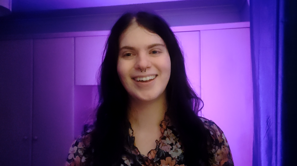 A smiling person with long dark hair and a septum piercing stands facing the camera, wearing a floral top. The background is softly lit in purple tones, with panelled walls creating a calm, studio-like atmosphere.