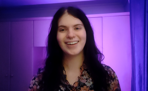 A smiling person with long dark hair and a septum piercing stands facing the camera, wearing a floral top. The background is softly lit in purple tones, with panelled walls creating a calm, studio-like atmosphere.
