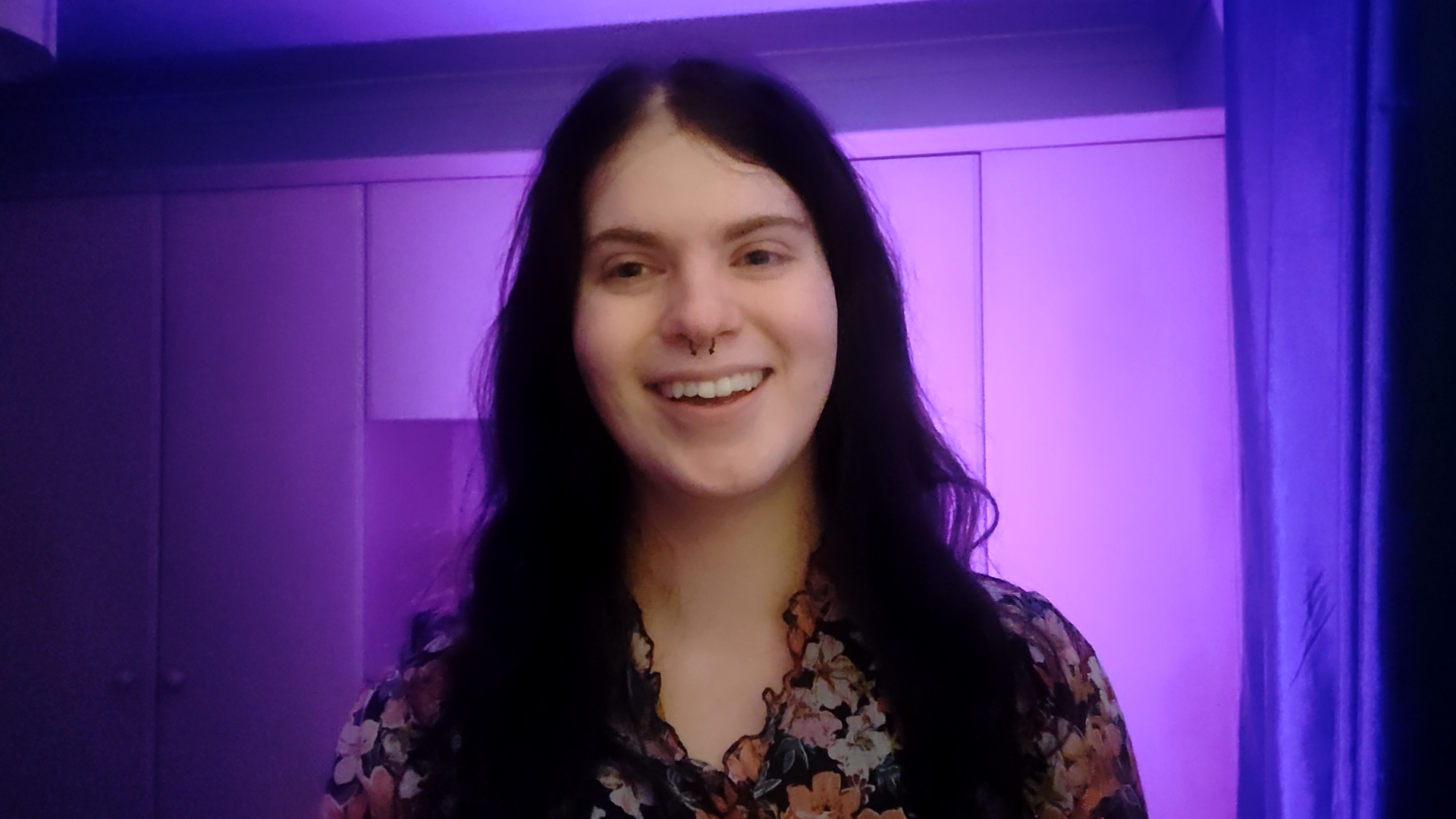 A smiling person with long dark hair and a septum piercing stands facing the camera, wearing a floral top. The background is softly lit in purple tones, with panelled walls creating a calm, studio-like atmosphere.